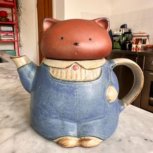 Op de afbeelding: Een keramische theepot in de vorm van een kat die een blauwe tuinbroek draagt. De kat heeft een bruine kop met eenvoudige zwarte ogen en een neus. Het lichaam is blauw met een crèmekleurige kraag, een klein roze knoopje, een tuit links en een handvat rechts.