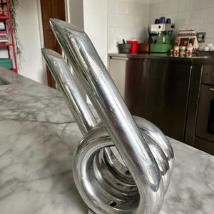 May include: A polished silver-toned metal sculpture featuring a coiled, circular base with two elongated, upward-pointing extensions. The object has a smooth, reflective surface and is mounted on a small, rectangular metal base.