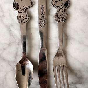 Può includere: Un set di posate in acciaio inossidabile da tre pezzi composto da un cucchiaio, un coltello e una forchetta. Il cucchiaio e la forchetta presentano un design con il personaggio Snoopy sul manico, mentre il manico del coltello mostra una grafica verticale di Snoopy con Woodstock.