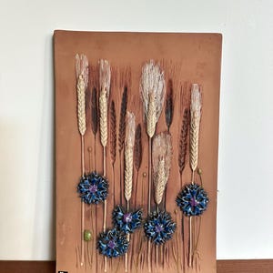 Puede incluir: Placa de pared rectangular de color terracota con un diseño en relieve de espigas de trigo y acianos azules. El trigo es de color beige claro, y las flores tienen pétalos azul oscuro con centros morados. La placa tiene una superficie texturizada.