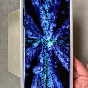 Può includere: Opera d'arte da parete rettangolare con un vivace design astratto blu e nero. L'opera è incorniciata in una cornice di metallo argentato e montata su un supporto in legno chiaro. Il design blu ha una trama maculata.