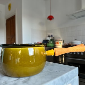 Könnte beinhalten: Ein gelb-grüner Keramik-Kochtopf mit Holzgriff. Der Topf hat einen schwarzen Rand und steht auf einer Marmorarbeitsplatte. Der Griff ist mit einem gelben Verbinder befestigt.