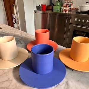 May include: Four ceramic cup and saucer sets in matte finishes. The colors include a pale cream, a vibrant coral, a deep blue, and a warm orange. Each set features a cylindrical cup with a flat-bottomed saucer, arranged on a marble surface.