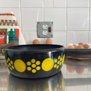 Può includere: Una casseruola smaltata nera con un motivo a pois gialli e un lungo manico. La scena della cucina comprende una tazza verde, uova e un canovaccio fantasia. La padella misura circa 20 cm di diametro.