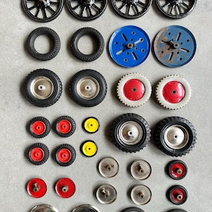 以下が含まれることがあります： さまざまなサイズとデザインの玩具の車輪の詰め合わせ。 黒いスポークホイール、ゴムタイヤ、青と赤のディスクホイール、赤または黄色のセンターが付いた小さなホイールが含まれています。 車輪は灰色の表面に配置されています。