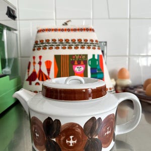 Könnte beinhalten: Weiße Keramik-Teekanne mit braunem Deckel und Griff, verziert mit braunen Blumenmustern. Die Teekanne steht auf einer Edelstahl-Oberfläche, mit einem gemusterten Küchentuch im Hintergrund.