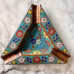 May include: A triangular cloisonné ashtray featuring a vibrant turquoise background with multi-colored floral patterns in red, pink, yellow, and blue. The interior base displays a geometric fretwork design, and the edges are finished in copper.