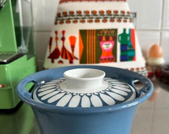 May include: A blue ceramic casserole dish with a white and blue floral pattern on the lid. The dish has two handles and a white knob. The background includes a patterned apron and a green appliance.