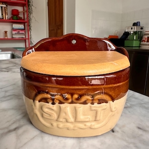 Pode incluir: Um saleiro de parede em cerâmica com acabamento vidrado em dois tons, castanho e bege, com a palavra SALT em relevo na parte frontal. O recipiente possui uma tampa plana de madeira clara e um painel traseiro curvo com um orifício de fixação.