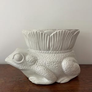 Pode incluir: Um vaso de cerâmica branca em forma de sapo, com um corpo texturizado e um topo em forma de lótus. O sapo tem olhos grandes e redondos e está sobre uma superfície de madeira. Um item decorativo.