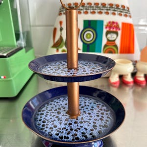 May include: Two-tiered serving stand with a copper-coloured central post and handle. The two circular trays are navy blue with a light blue and white patterned surface. The stand is designed for displaying or serving food items.
