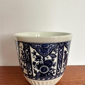 Pode incluir: Um vaso de cerâmica com borda e base creme, e um padrão floral azul e branco. O vaso tem formato cilíndrico com a parte superior ligeiramente alargada e uma base canelada. O design floral inclui flores e folhas estilizadas.