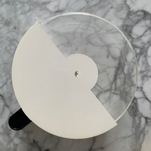 Pode incluir: Um recipiente de armazenamento de alimentos de plástico branco com uma tampa transparente. A tampa tem um pequeno furo no centro.