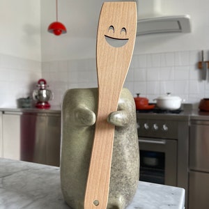 Puede incluir: Una espátula de madera con un diseño de cara sonriente es sostenida por un soporte de cerámica para utensilios. La espátula es de color marrón claro y el soporte es de color gris verdoso moteado. El entorno de la cocina incluye electrodomésticos de acero inoxidable y una lámpara colgante roja.