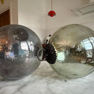 Op de afbeelding: Twee grote, ronde kerstballen. Eén is zilverkleurig, de andere is lichtgroen. Beide hebben geëtste sterpatronen. Elke bal heeft een donkerbruine dop met een metalen ring om op te hangen. De ballen liggen op een marmeren oppervlak.