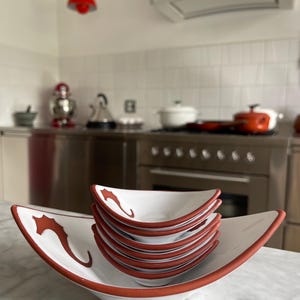 Könnte beinhalten: Ein Set aus sieben weißen Keramikschalen mit rotem Rand und einem Seepferdchen-Design. Die Schalen sind übereinander gestapelt und stehen auf einer weißen Marmor-Arbeitsplatte.