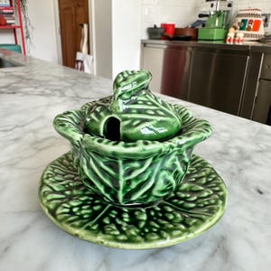 May include: A green ceramic cabbage-shaped serving dish with a lid and a matching saucer. The dish has a glossy finish and detailed leaf patterns. The lid features a small handle and a hole for a spoon.