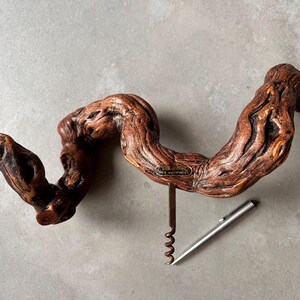 Könnte beinhalten: Ein Korkenzieher mit einem Griff aus einem knorrigen, dunkelbraunen Stück Rebholz. Der Griff hat ein kleines Metallschild mit der Aufschrift 'Cépages des vignes'. Eine silberfarbene Metallspirale und ein Hebel sind am Holz befestigt.