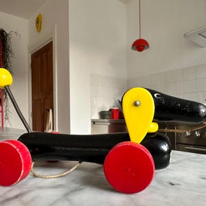 Può includere: Un giocattolo vintage in legno a forma di cane da tirare. Il cane è nero con ruote rosse e una testa e pinna caudale gialle. Ha una palla gialla su una molla e una corda per tirare.
