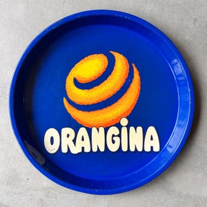 Pode incluir: Uma bandeja de servir redonda de metal azul profundo com o logótipo laranja em forma de espiral da Orangina no centro. Abaixo do gráfico, a palavra ORANGINA está impressa em letras grandes, a negrito e de cor creme. A bandeja tem uma borda elevada.