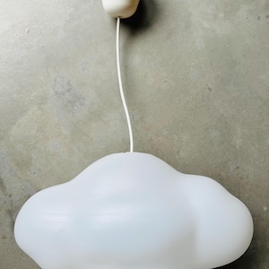 May include: A white cloud-shaped pendant light fixture. The light has a rounded, irregular shape and hangs from a white cord and a white ceiling mount. The fixture is held by a hand.