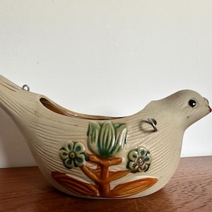 May include: A beige ceramic bird-shaped planter with a textured finish and floral decorations in green and brown. It has a planting opening and a metal loop for hanging. The planter is designed to be wall-mounted.