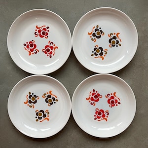 Könnte beinhalten: Vier weiße Keramikplatten mit floralem Design. Jede Platte zeigt drei stilisierte Blumen in Rot-, Orange- und Brauntönen. Die Teller sind quadratisch angeordnet und präsentieren das passende Design.