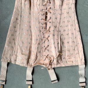 Puede incluir: Un corsé melocotón vintage con un estampado floral y cierre de cordones en la parte delantera. El corsé tiene correas ajustables y sujeta ligas. La tela tiene un brillo sutil y el diseño es simétrico.