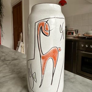 Könnte beinhalten: Eine hohe, weiße Keramikvase mit zylindrischer Form. Die Vase zeigt eine stilisierte orangefarbene und schwarze Illustration eines Tieres. Schwarze Linien umreißen das Tier und andere abstrakte Formen. Die Vase hat eine strukturierte Oberfläche und einen braunen Rand.