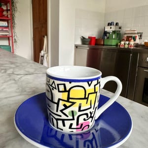 Könnte beinhalten: Eine weiße Kaffeetasse aus Keramik mit passender Untertasse. Die Tasse ist mit einem markanten schwarzen, abstrakten Linienmuster auf gelben, rosafarbenen und grünen Formen verziert. Die Untertasse und der Tassenrand sind in einem kräftigen Blau.