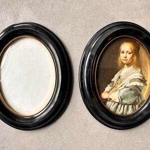 May include: Two oval-shaped picture frames with glossy black finishes. One frame is empty with a white backing, while the other contains a portrait painting of a person with long blonde hair wearing a light-colored collared garment and a blue jacket.
