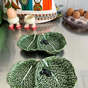 May include: Two green ceramic leaf-shaped dishes with a textured surface and dark olives. The dishes are on a shiny metal surface. In the background are egg holders, eggs, and a patterned kitchen towel.