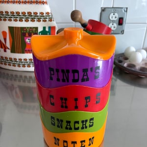 May include: A stack of four colourful plastic containers with lids. The containers are labelled "Pinda's", "Chips", "Snacks", and "Noten".