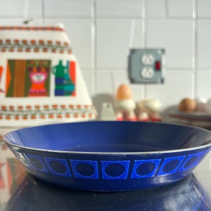 May include: A blue enamel serving bowl with a silver rim. The bowl has a repeating pattern of blue squares with oval centres. The bowl is sitting on a reflective surface.