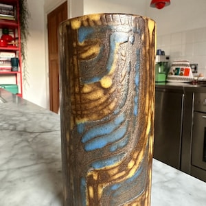 Puede incluir: Un jarrón cilíndrico de cerámica con una superficie texturizada. El jarrón presenta un patrón de remolinos marrones, amarillos y azules. Este jarrón decorativo es adecuado para exhibir flores o como pieza de arte independiente.