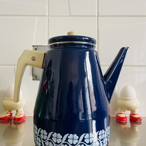 Könnte beinhalten: Eine blaue Emaille-Teekanne mit weißem Blumenmuster. Die Teekanne hat einen cremefarbenen Griff und Deckel.