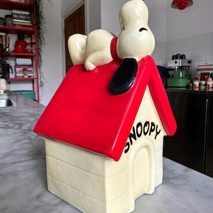 Peut inclure: Un pot à biscuits en céramique en forme de niche Snoopy. La niche a un toit rouge et un corps crème avec le mot "SNOOPY" en noir. Snoopy est allongé sur le toit, avec une oreille noire et un collier rouge.