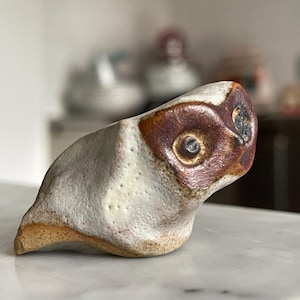 Może przedstawiać: Ceramiczna rzeźba sowy z brązową i białą glazurą. Sowa patrzy w prawo i ma fakturowaną powierzchnię.