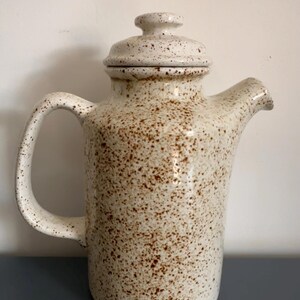 May include: A ceramic coffee pot with a speckled cream and brown finish. The pot features a cylindrical body, a curved handle on the left side, a short spout on the right, and a matching removable lid with a small knob handle on top.