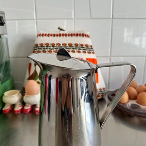 Puede incluir: Una jarra de acero inoxidable con tapa y asa. La jarra tiene una superficie lisa y pulida. El asa es rectangular y está fijada al lateral. Probablemente se utiliza para servir líquidos.
