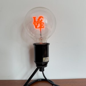 Peut inclure: Une ampoule décorative avec le mot "LOVE" en néon orange. L'ampoule est transparente et repose sur une base noire avec un fil et un support noirs. La base porte une étiquette indiquant "Silly max 40 w".