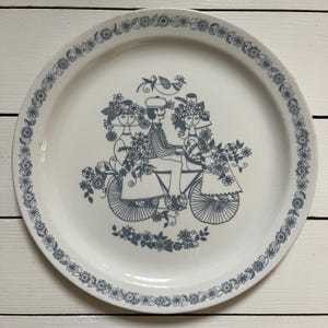 Może przedstawiać: Biały ceramiczny talerz z niebieską kwiatową obwódką i centralną ilustracją pary na rowerze, otoczonej kwiatami. Talerz ma wygląd vintage i nadaje się do serwowania potraw.