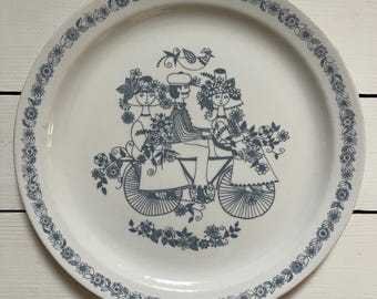 May include: White ceramic plate with a blue floral border and a central illustration of a couple riding a bicycle, surrounded by flowers. The plate has a vintage aesthetic and is suitable for serving food.