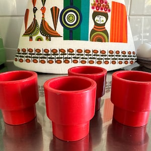 May include: Four bright red egg cups with a stepped design. The cups are arranged on a reflective surface, with a colorful patterned fabric in the background. The fabric features birds, flowers, and geometric shapes. A vintage kitchenware set.