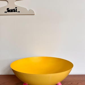 Può includere: Una ciotola giallo brillante con interno ed esterno lisci e arrotondati, sostenuta da tre piccoli piedini cilindrici rosa. La ciotola presenta un'apertura ampia e un design a bacino poco profondo e curvo, che offre una base stabile.