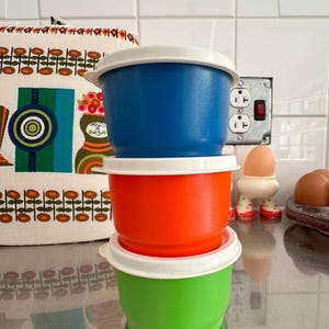 Könnte beinhalten: Drei Vintage-Kunststoff-Lebensmittelbehälter in Blau, Orange und Grün, jeweils mit einem weißen Deckel. Die Behälter sind übereinander gestapelt. Der Hintergrund umfasst einen gemusterten Stoff und eine weiße geflieste Wand.