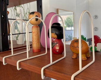 May include: White metal bookends with three dividers on a wooden shelf. The bookends are holding a variety of decorative items, including a pink egg-shaped object, a wooden doll with a red and white kimono, and a wooden dog figurine.