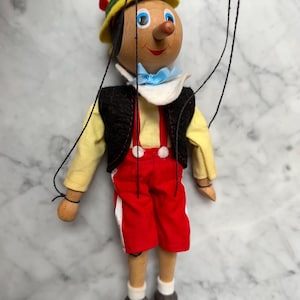 Puede incluir: Títere de madera de Pinocho, con sombrero amarillo, pantalones cortos rojos y chaleco negro. La marioneta tiene una nariz larga y está suspendida por hilos. Un juguete infantil clásico.