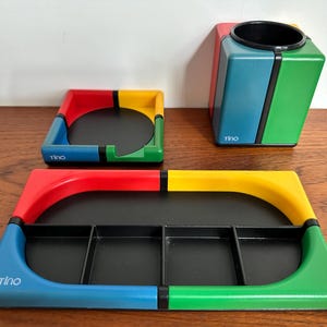 May include: A set of colorful desk organizers in primary colors: red, yellow, blue, and green. The set includes a pen holder, a small square coaster, and a rectangular tray with four compartments. The word "tino" is printed on each item.