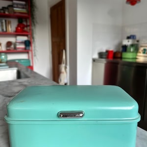 Peut inclure: Une boîte à pain vintage de couleur turquoise, de forme rectangulaire avec un couvercle incurvé. Elle est dotée d'une poignée argentée sur le devant. La boîte à pain est en métal et repose sur un comptoir en marbre. Environ 30 cm de long.